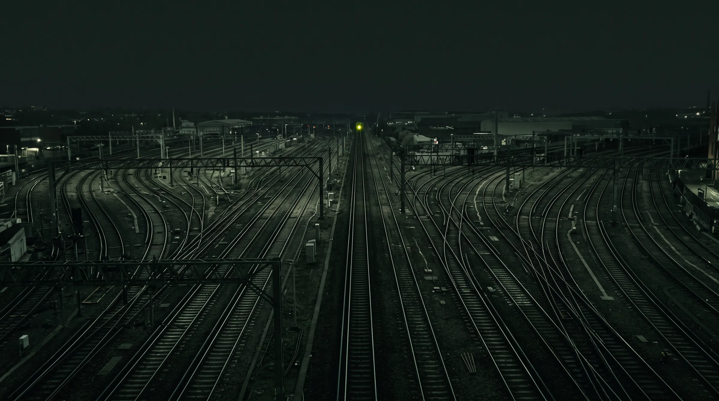 Railway tracks at night converging into a single main line under one green signal.